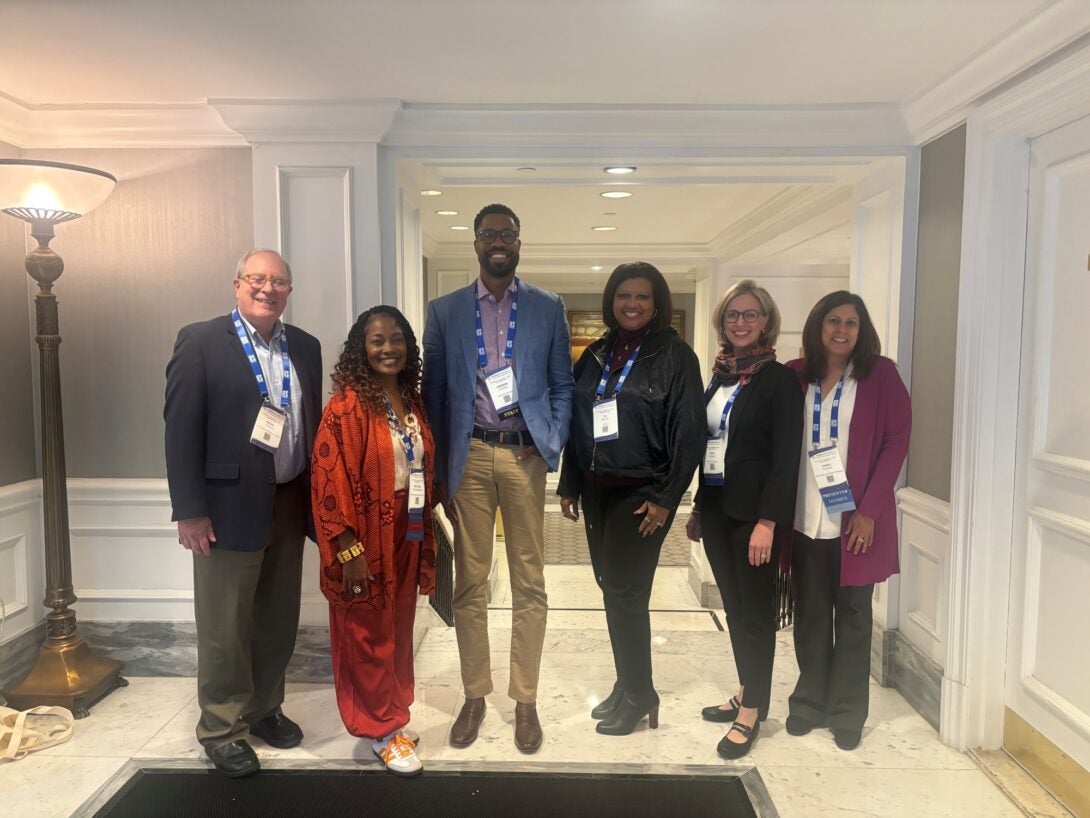 The photo features five people standing together in a row, smiling and all wearing white conference badges with blue lanyards around their necks. The location is a hallway with tan walls, white doors and paneling, bright lights, and white marble floors. An elegant bronze standing lamp is right behind and to the right of them, in the left hand edge of the frame.