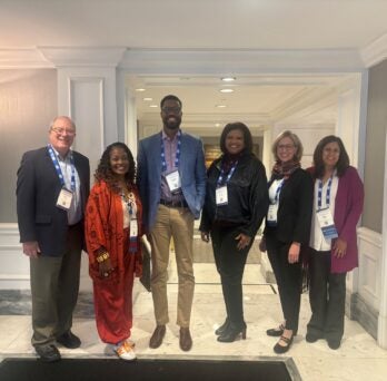 The photo features five people standing together in a row, smiling and all wearing white conference badges with blue lanyards around their necks. The location is a hallway with tan walls, white doors and paneling, bright lights, and white marble floors. An elegant bronze standing lamp is right behind and to the right of them, in the left hand edge of the frame. 