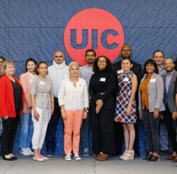 The photo features two rows of people, with 11 people standing in the back row and 12 people standing in the front row. They are standing against a blue textured wall with a large red circle in the center and the outline of the letters “UIC” cut out in the middle of the red circle. 