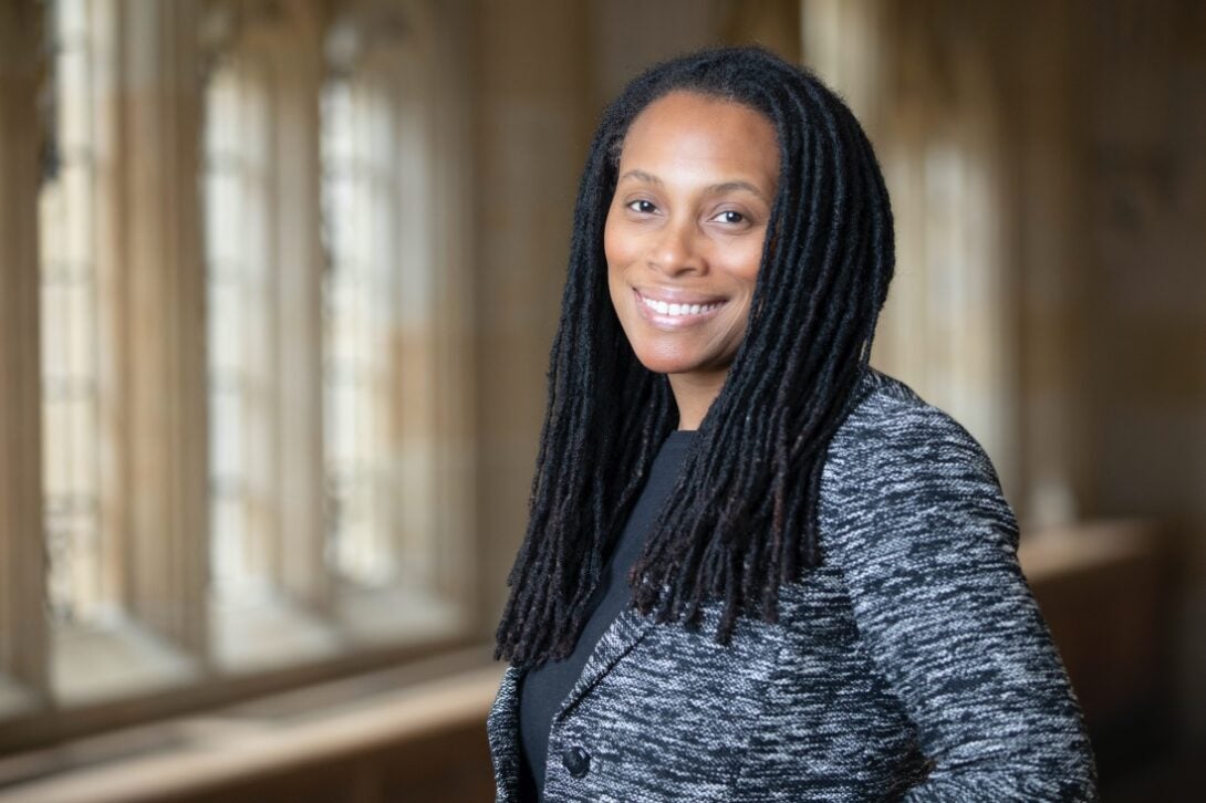 The photo features a person with long, dark dreadlocks smiling at the camera. She is angled slightly away from the camera and is wearing a dark grey shirt with a blazer over it; the blazer has a dark and light grey flamestitch pattern. There are gothic style windows behind her with sunlight pouring in, illuminating the space. The background is softly blurred to highlight the person in the frame.