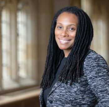 The photo features a person with long, dark dreadlocks smiling at the camera. She is angled slightly away from the camera and is wearing a dark grey shirt with a blazer over it; the blazer has a dark and light grey flamestitch pattern. There are gothic style windows behind her with sunlight pouring in, illuminating the space. The background is softly blurred to highlight the person in the frame. 